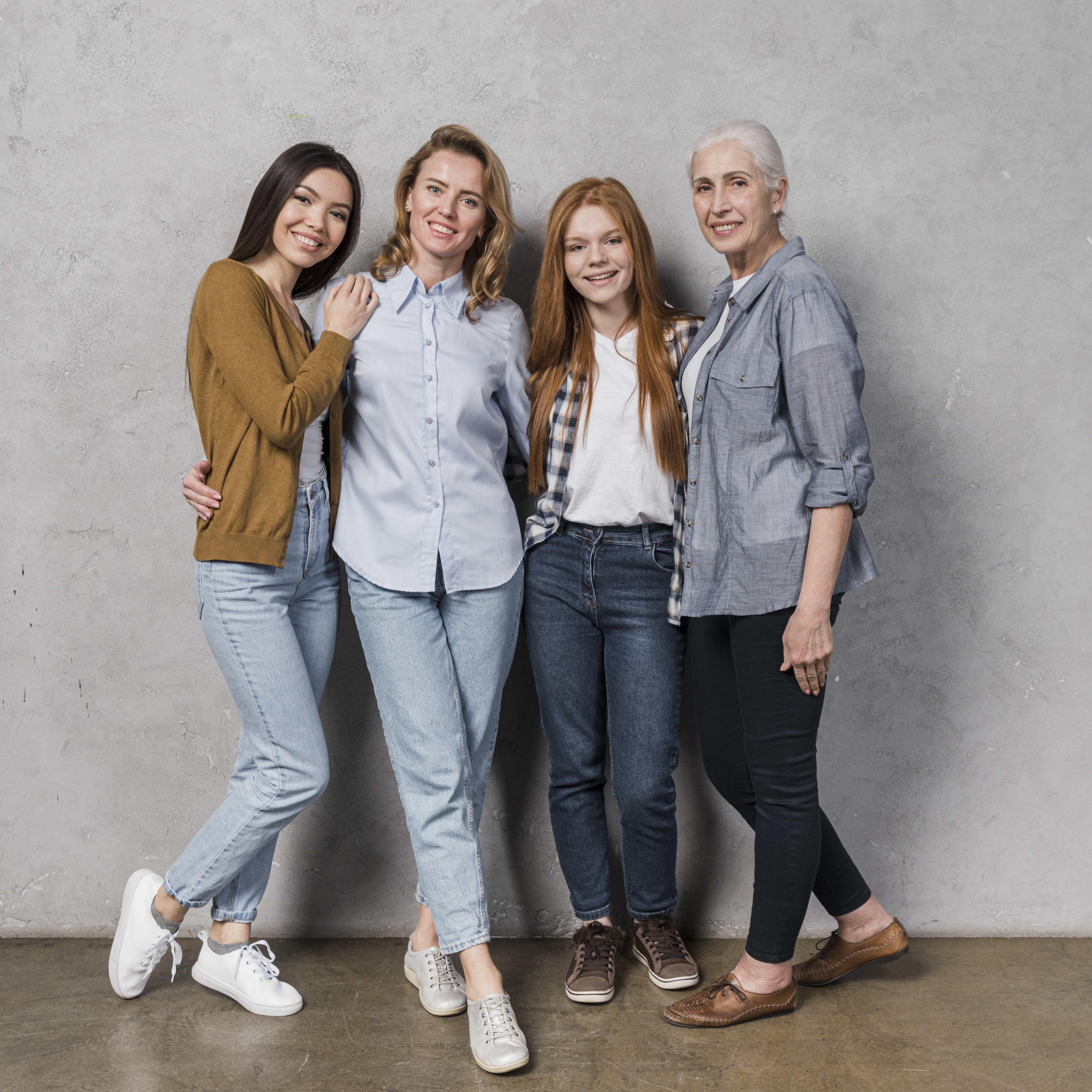 Quatre femmes de différents âges posent ensemble, souriantes, devant un mur gris.